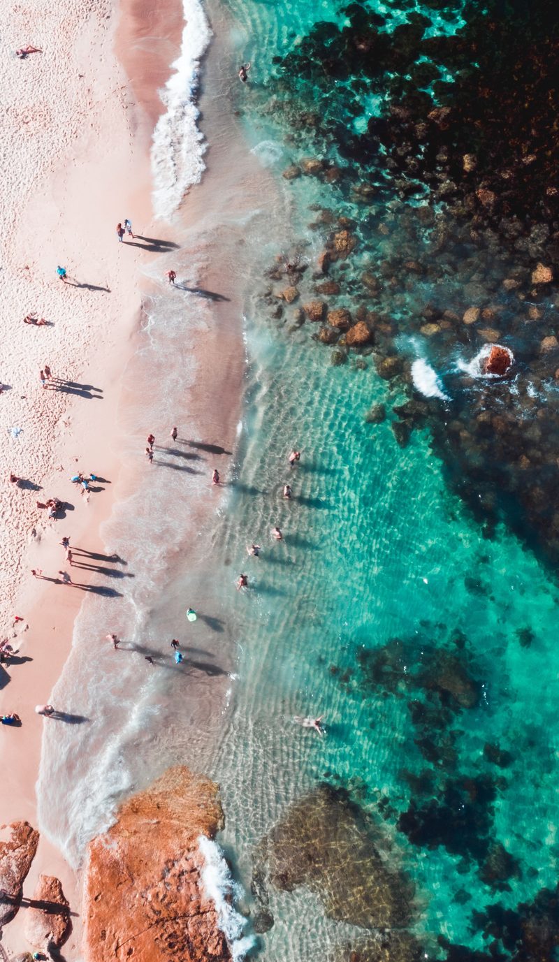 Swimmers and beachgoers on the sand and in the clear turquoise water, seen from above