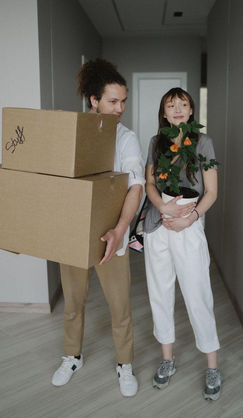 A couple carrying moving boxes and a pot plant into their first home
