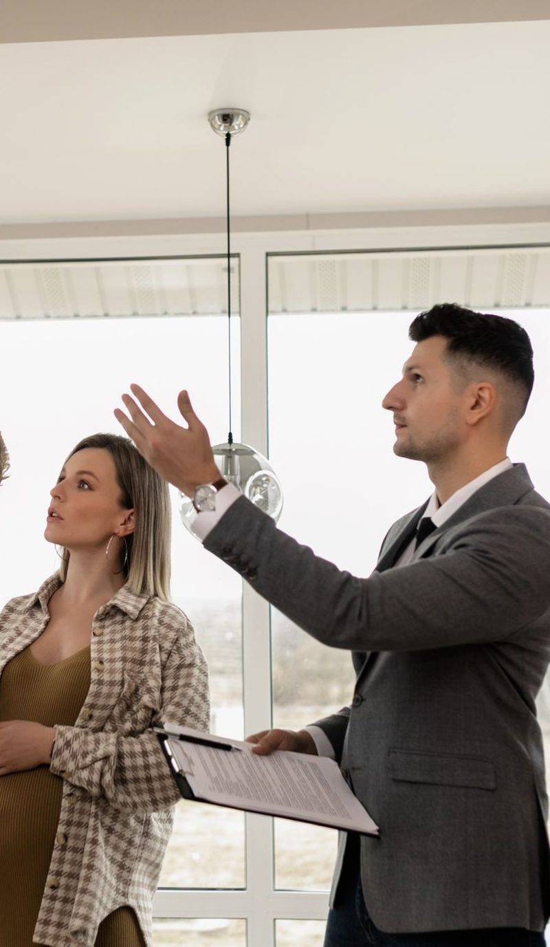 An agent showing a couple through a bright empty room with floor to ceiling windows at an inspection