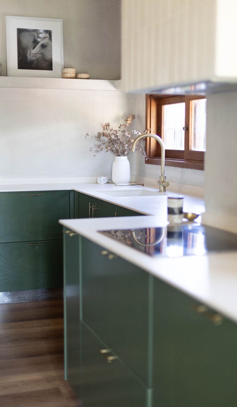 A renovated kitchen with dark green cabinetry, a white stone benchtop, a brass tap over the sink and a timber framed window