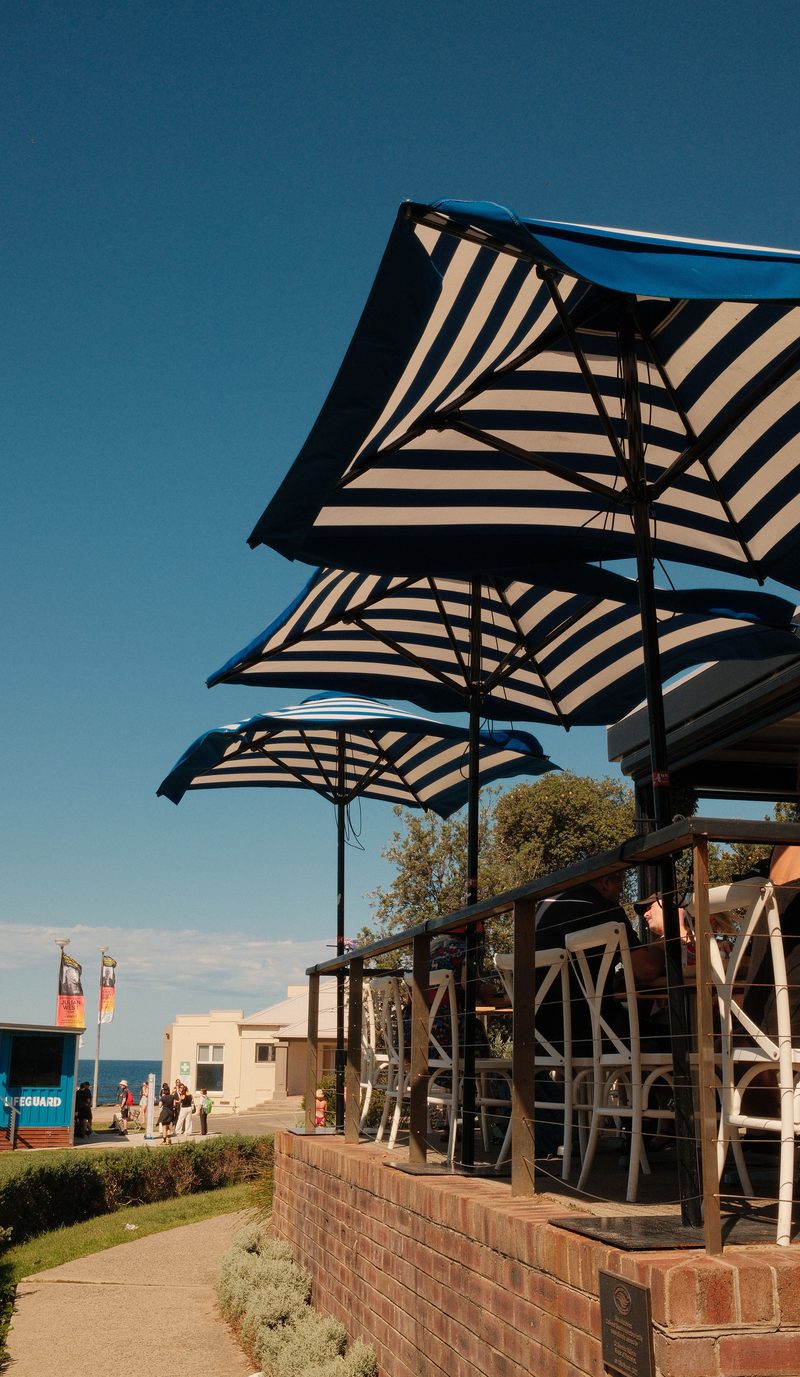 Striped umbrellas over the terrace of a beachfront cafe, with the ocean and a lifeguard tower beyond