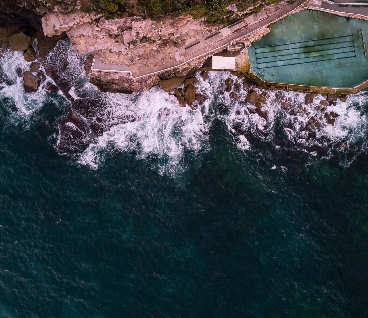 An ocean pool cut into the rocks below the cliffs, with surf breaking around it, seen from above
